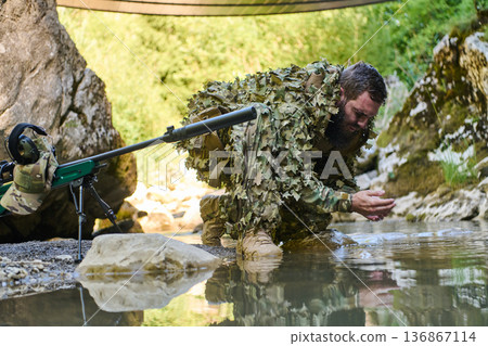 Sniper In Ghillie Suit Kneeling By Creek In Camouflage Doing Water Reconnaissance Outdoors 136867114