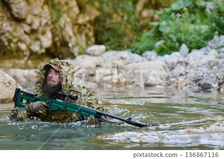 Camouflaged Sniper Soldier Wading Through River With Rifle in Woodland Ghillie Suit Camouflaged Sniper Soldier Wading Through River With Rifle in Woodland Ghillie Suit 136867116