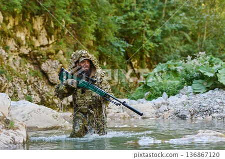 Camouflaged Sniper Soldier Wading Through River In Ghillie Suit During Recon Mission In Forest 136867120