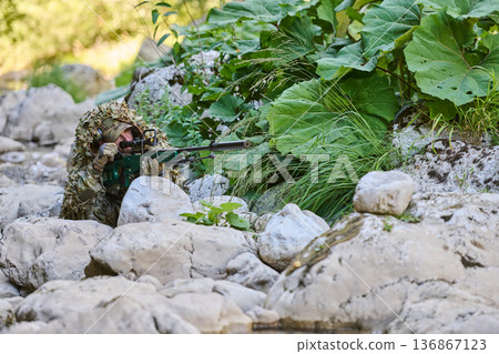 Camouflaged Sniper Soldier Lying Among Rocks and Foliage With Scoped Rifle in Concealment 136867123