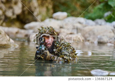 Camouflaged Sniper in Ghillie Suit Aiming Rifle While Partially Submerged in River 136867124