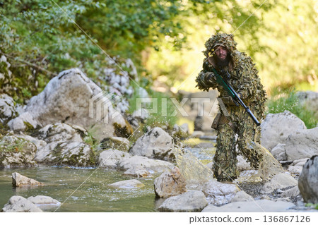 Soldier In Ghillie Suit Moving Through Rocky Riverbed With Rifle Tactical Camouflage Soldier In Ghillie Suit Moving Through Rocky Riverbed With Rifle Tactical Camouflage 136867126