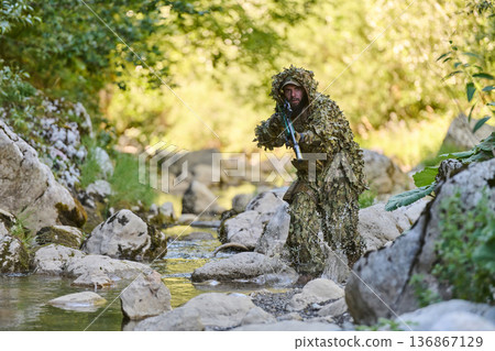Camouflaged Soldier In Ghillie Suit Moving Through Rocky Stream With Rifle On Recon Mission 136867129
