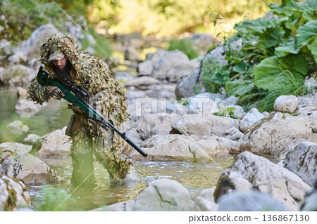 Camouflaged Sniper In Ghillie Suit Crossing Rocky Stream During Military Reconnaissance In Woodland 136867130