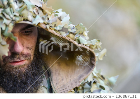Close-Up Of Camouflaged Sniper In Ghillie Suit With Face Paint And Beard Outdoors 136867131