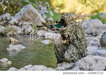 Camouflaged Sniper In Ghillie Suit Aiming A Suppressed Rifle While Kneeling By Rocky River Camouflaged Sniper In Ghillie Suit Aiming A Suppressed Rifle While Kneeling By Rocky River 136867132