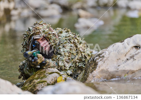 Sniper In Ghillie Suit Camouflaged On Riverbank Aiming Tactical Rifle In Stealth Operation Sniper In Ghillie Suit Camouflaged On Riverbank Aiming Tactical Rifle In Stealth Operation 136867134