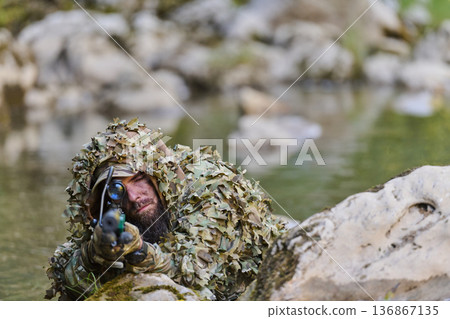 Sniper in Ghillie Suit Aiming Through Scope While Concealed on Rocky Creek Bank 136867135