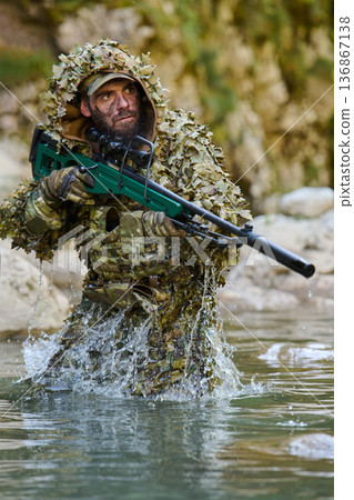 Sniper Soldier in Ghillie Suit Wading Through River With Rifle in Tactical Camouflage 136867138