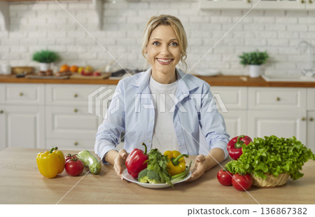 Portrait of happy smiling young woman sitting at kitchen table with fresh vegetables 136867382