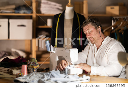 Man seamstress operating by sewing machine, manufactures clothing 136867395