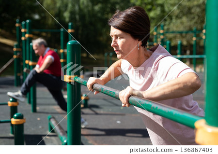 elderly woman performs exercises on horizontal bars in an outdoor sports area 136867403