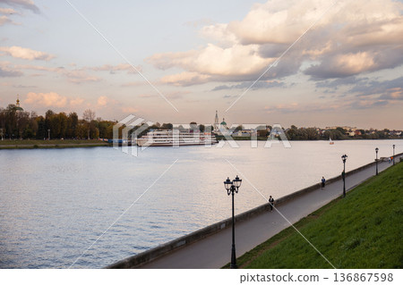 Riverside walkway with lampposts along calm river and distant church under cloudy sky 136867598