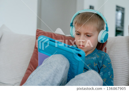 Little boy wearing wireless headphones sitting comfortably on sofa at home and playing on tablet. Cozy interior, modern technology and everyday digital entertainment concept. Little boy wearing wireless headphones sitting comfortably on sofa at home and playing on tablet. Cozy interior, modern technology and everyday digital entertainment concept. 136867905