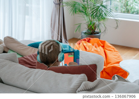 Child focused on tablet screen while relaxing indoors. Home atmosphere, safe environment and modern screen time activity for kids. Concept of digital childhood. 136867906