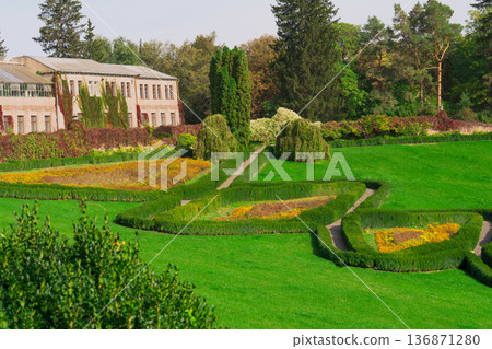 Green manicured lawn in the park, along which paths stretch, shrouded on both sides by cultivated shrubs and flowers, and extend to the house, behind which there are forest trees 136871280