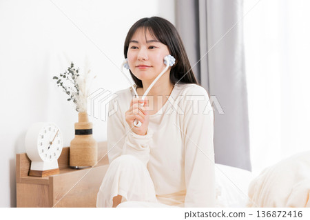 一位穿著睡衣的年輕亞洲女性正在使用臉部按摩器進行按摩。 136872416