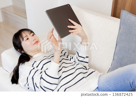 一名女子躺在沙發上看著平板電腦。 一名女子躺在沙發上看著平板電腦。 136872425