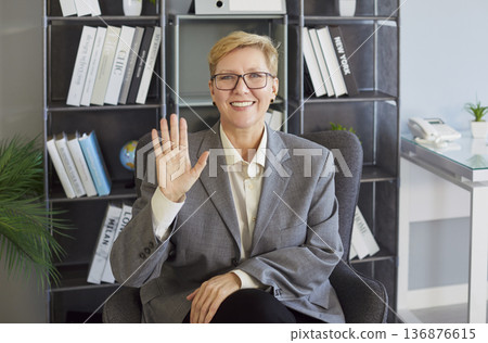 Smiling business professional waving hello during video call in modern office workspace 136876615