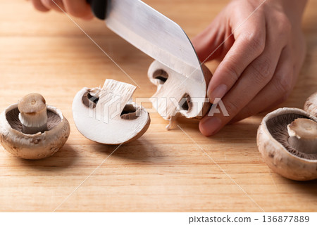 Hands holding kitchen knife and cutting cremini mushrooms on a wooden board Hands holding kitchen knife and cutting cremini mushrooms on a wooden board 136877889
