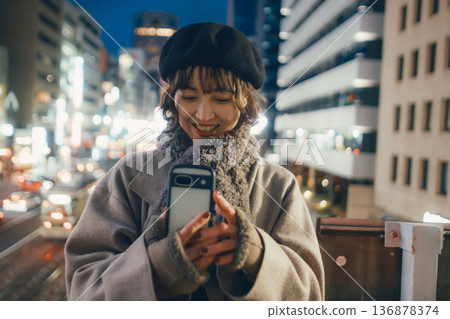 一位微笑的日本女子在夜晚的城市使用智慧型手機。 136878374