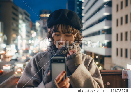 一位微笑的日本女子在夜晚的城市使用智慧型手機。 一位微笑的日本女子在夜晚的城市使用智慧型手機。 136878376