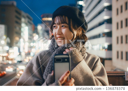 一位微笑的日本女子在夜晚的城市使用智慧型手機。 136878378