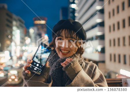 一位微笑的日本女子在夜晚的城市使用智慧型手機。 136878380