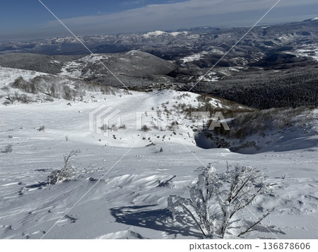 Snow-covered mountain slope overlooking a winter valley with distant peaks under a blue sky 136878606