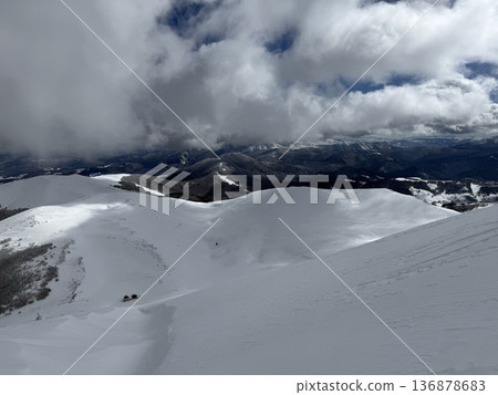 Snow-covered mountain ridge under dramatic clouds with distant valley and solitary cabin 136878683