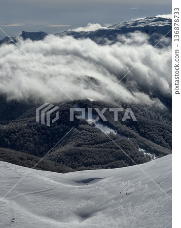 Snowy mountain ridge with low clouds rolling over forested valley in winter landscape Snowy mountain ridge with low clouds rolling over forested valley in winter landscape 136878733