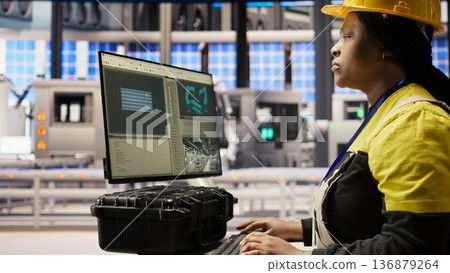 Woman in smart factory expert uses computer to implement automated workflows. African american technician using PC in industry 4.0 plant to integrate sensors into production, camera A Woman in smart factory expert uses computer to implement automated workflows. African american technician using PC in industry 4.0 plant to integrate sensors into production, camera A 136879264