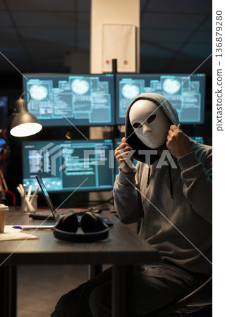 Anonymous man adjusting mask and hoodie while sitting at desk surrounded by glowing system monitors. Programmer with concealed identity seated in dark office room, planning to steal sensitive data. 136879280
