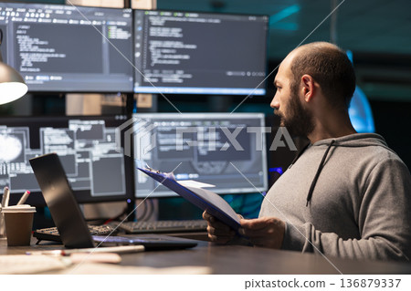 Programmer examines paperwork with serious expression and compares notes to code displayed on surrounding monitors. Developer with clipboard glancing at large displays of data, ensuring accuracy. Programmer examines paperwork with serious expression and compares notes to code displayed on surrounding monitors. Developer with clipboard glancing at large displays of data, ensuring accuracy. 136879337