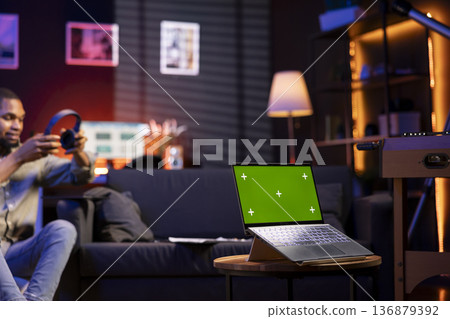 Young freelancer enjoys leisure time next to chroma key on the bean bag, uses his smartphone to watch a podcast with headset on. Black person relaxing remotely in the living room. Young freelancer enjoys leisure time next to chroma key on the bean bag, uses his smartphone to watch a podcast with headset on. Black person relaxing remotely in the living room. 136879392