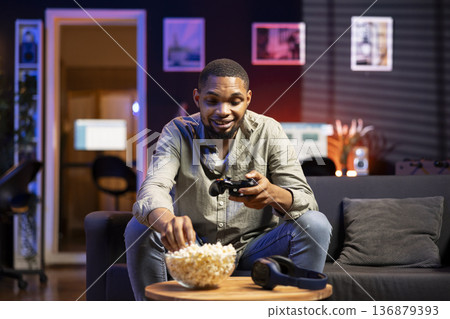 Young guy having fun with video games on television modern setup, connecting his console on smart tv in living room and enjoying chill game night at home. African american gamer playing tournament. Young guy having fun with video games on television modern setup, connecting his console on smart tv in living room and enjoying chill game night at home. African american gamer playing tournament. 136879393