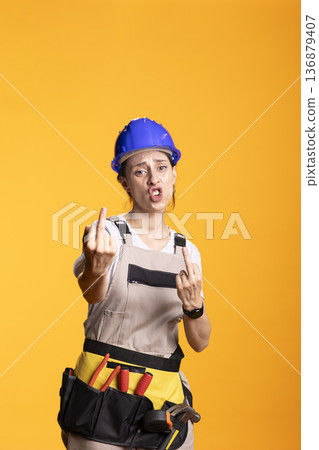 Female construction worker displays bold expressive gesture while wearing safety helmet and toolbelt. Studio portrait communicates strong emotion, attitude or reaction in professional work context. Female construction worker displays bold expressive gesture while wearing safety helmet and toolbelt. Studio portrait communicates strong emotion, attitude or reaction in professional work context. 136879407