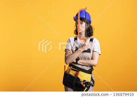 Female renovation worker poses with hacksaw tool, standing with arms crossed and playful facial expression. Confident repairwoman wearing helmet holds hacksaw in front of eye for humorous expression. 136879410