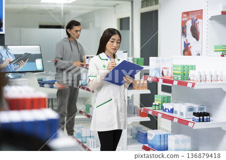 Pharmacy expert inspects packaged medications on shelves, managing inventory in modern drugstore. Asian pharmacist stands in dispensary aisle with clipboard, checking health products availability. 136879418