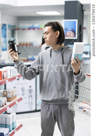Asian man with shopping basket inspects health supplements in neighborhood pharmacy. Male customer evaluates pharmaceutical products carefully, emphasizing responsible personal health and selfcare. Asian man with shopping basket inspects health supplements in neighborhood pharmacy. Male customer evaluates pharmaceutical products carefully, emphasizing responsible personal health and selfcare. 136879419