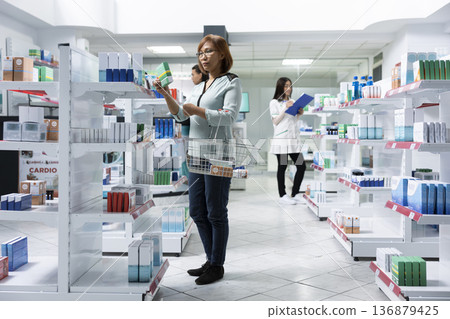 Middle aged woman browsing pharmacy shelves, selecting medication to support her health and wellbeing. Asian female customer shops for wellness supplements in a neighborhood dispensary. Middle aged woman browsing pharmacy shelves, selecting medication to support her health and wellbeing. Asian female customer shops for wellness supplements in a neighborhood dispensary. 136879425
