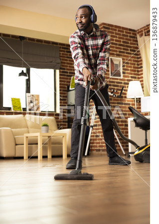 African American man enjoying music through headphones while vacuuming living room floor, completing weekend chores. Relaxed black male individual tidying up apartment with vacuum on sunny afternoon. African American man enjoying music through headphones while vacuuming living room floor, completing weekend chores. Relaxed black male individual tidying up apartment with vacuum on sunny afternoon. 136879433
