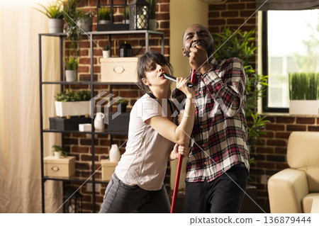 Happy white woman and black man singing into mop and TV remote like microphones, having during cleaning day. Diverse couple enjoying domestic chores, turning home cleanup into joyful activity. Happy white woman and black man singing into mop and TV remote like microphones, having during cleaning day. Diverse couple enjoying domestic chores, turning home cleanup into joyful activity. 136879444