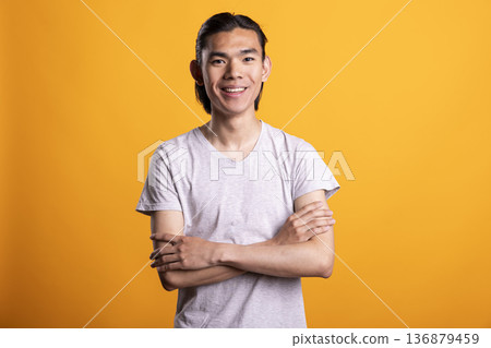 Smiling young Asian man stands confidently with folded arms, showing happiness and fulfillment. Proud and satisfied male model poses, radiating positive attitude and cheerful self assurance. 136879459