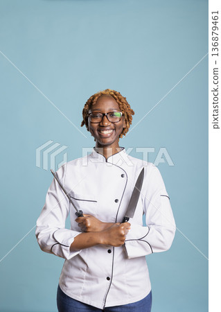 Cheerful black woman standing with knife and sharpener in hand, ready to begin culinary journey. Professional cook poses confidently with kitchen tools, expressing skill and enthusiasm for cooking. Cheerful black woman standing with knife and sharpener in hand, ready to begin culinary journey. Professional cook poses confidently with kitchen tools, expressing skill and enthusiasm for cooking. 136879461