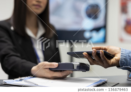 Black person completes cashless transaction at private clinic using smartphone and digital wallet. Asian female receptionist handles medical billing process with pos as patient taps with mobile device Black person completes cashless transaction at private clinic using smartphone and digital wallet. Asian female receptionist handles medical billing process with pos as patient taps with mobile device 136879463