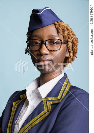 Professional portrait of African American flight attendant wearing formal aviation uniform. Closeup of air hostess looking at camera with proud and determined expression against blue background. 136879464