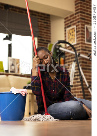 African american woman sits on floor, leaning on mop handle with eyes closed, looking tired after daily cleaning chores. Weary housewife takes break from mopping in brick wall living room. African american woman sits on floor, leaning on mop handle with eyes closed, looking tired after daily cleaning chores. Weary housewife takes break from mopping in brick wall living room. 136879487
