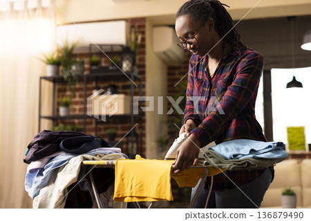 Young african american woman in casual outfit carefully pressing wrinkled clothes with electric iron on ironing board. Focused female individual doing household chores in cozy modern apartment. 136879490
