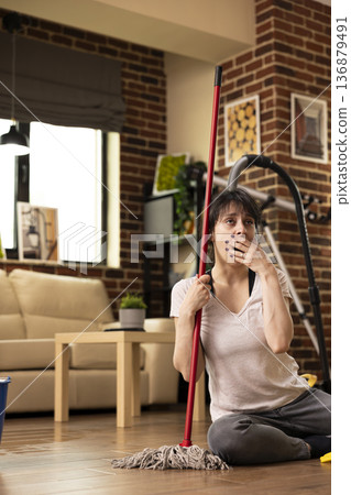 Caucasian woman sits on floor with mopping stick, yawning and covering her mouth, exhausted after long day of housecleaning. Young lady takes moment to rest before completing household chores. 136879491